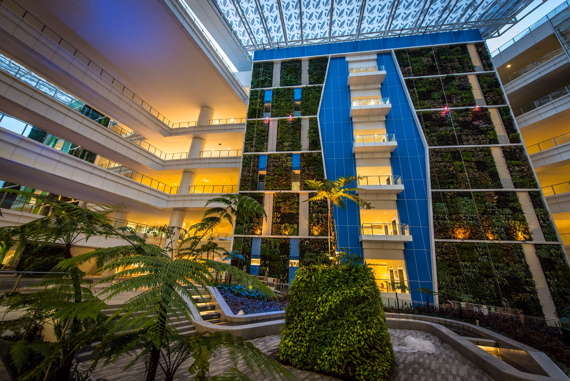 Multi-storey atrium at dusk with a lit green wall, warm-lit balconies and a patterned canopy roof