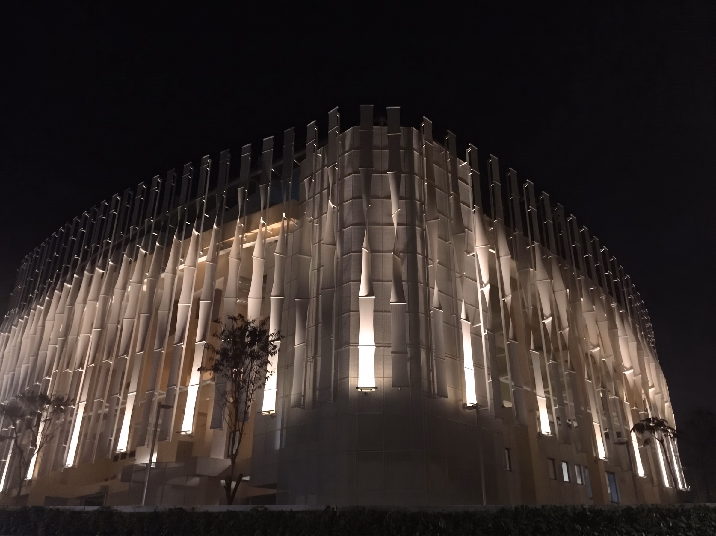 Civic building at night with vertical fins on a curved facade, each grazed from below by warm light