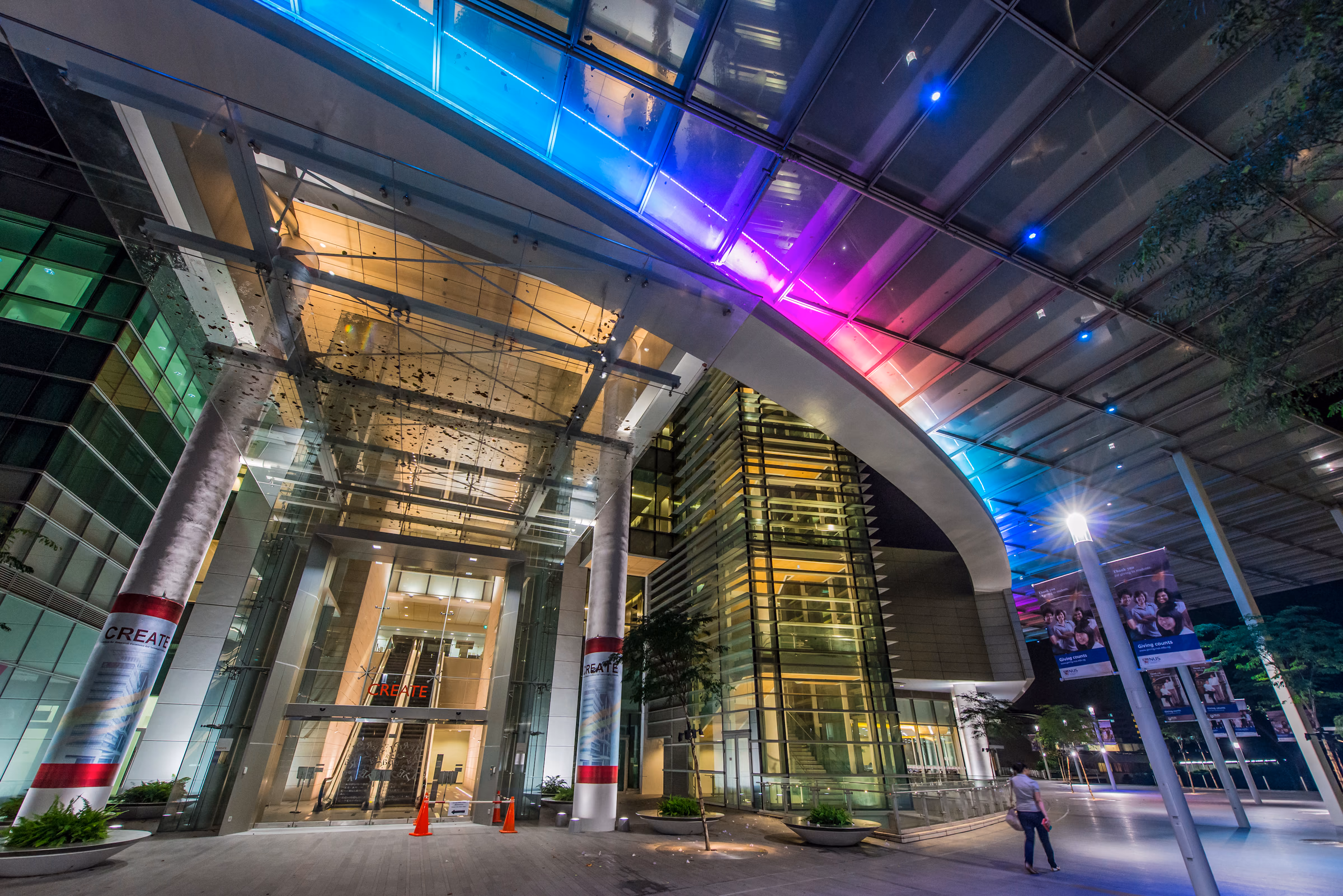 Glass campus entrance at night under a canopy lit in bands of blue, magenta and cyan, with warm light spilling from the atrium behind