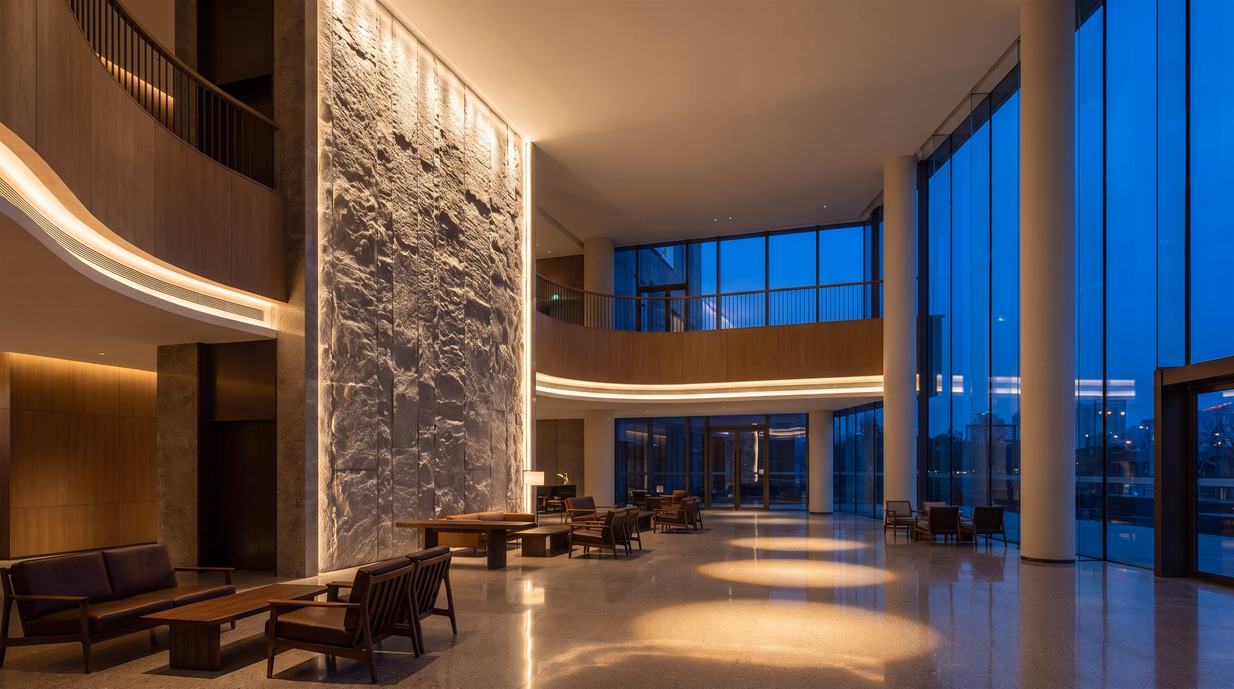 Double-height lobby atrium at dusk with a grazed stone wall and concealed cove lighting along the balconies