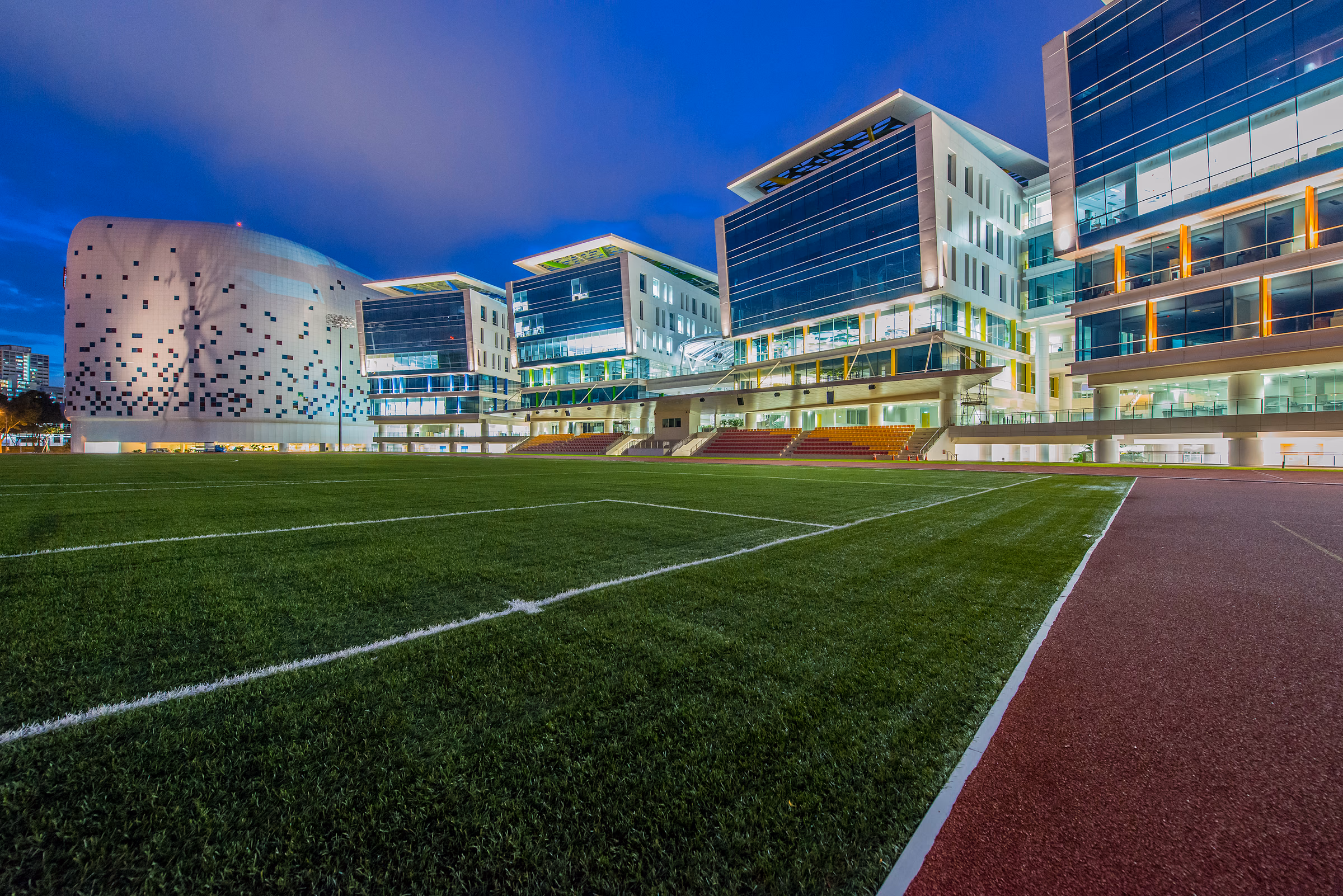 Campus sports field at dusk with the lit teaching blocks and a perforated drum-shaped hall behind it