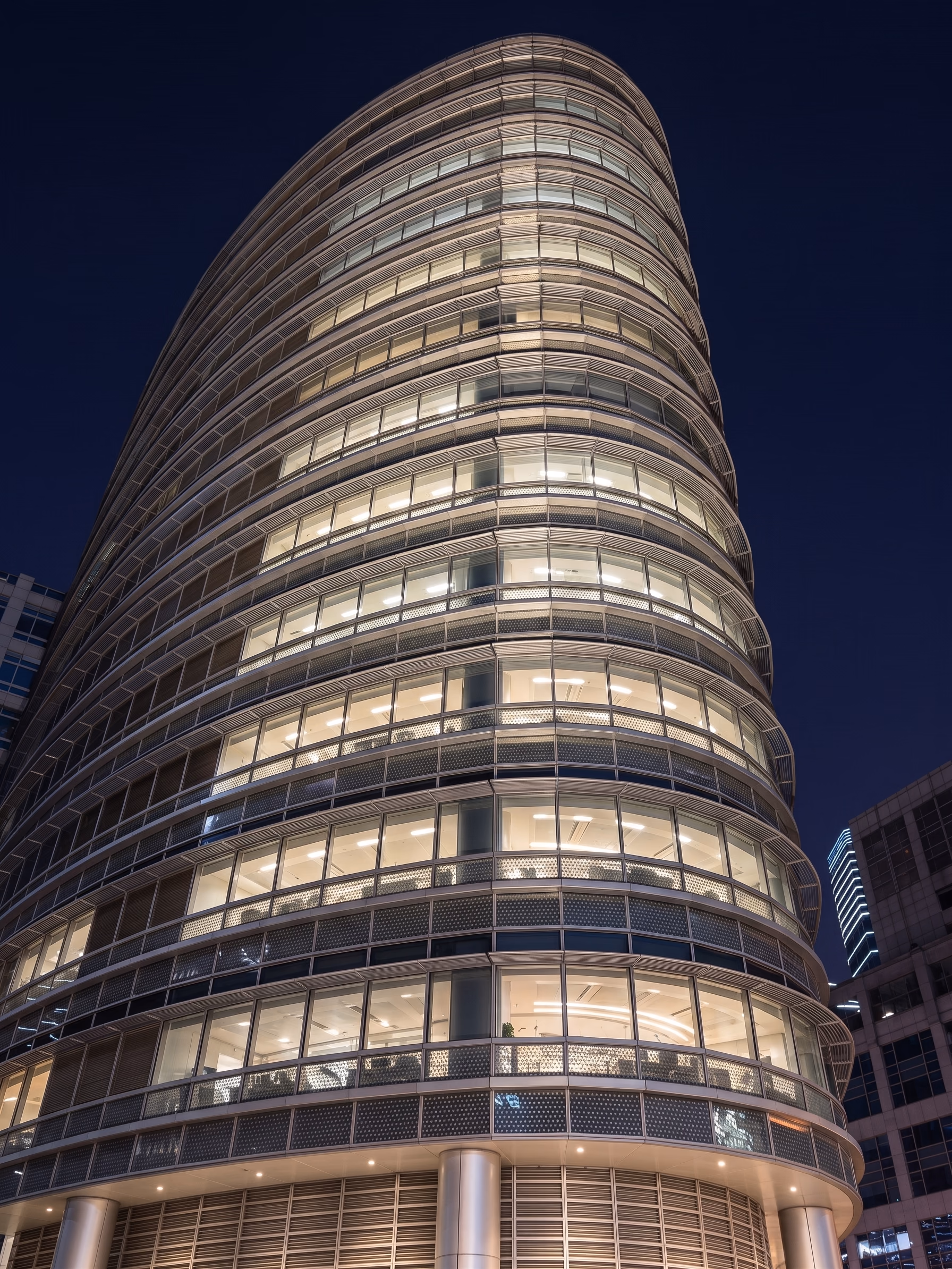 Curved office tower at night seen from below, its floor plates glowing behind glazing with a lit band at every balustrade line