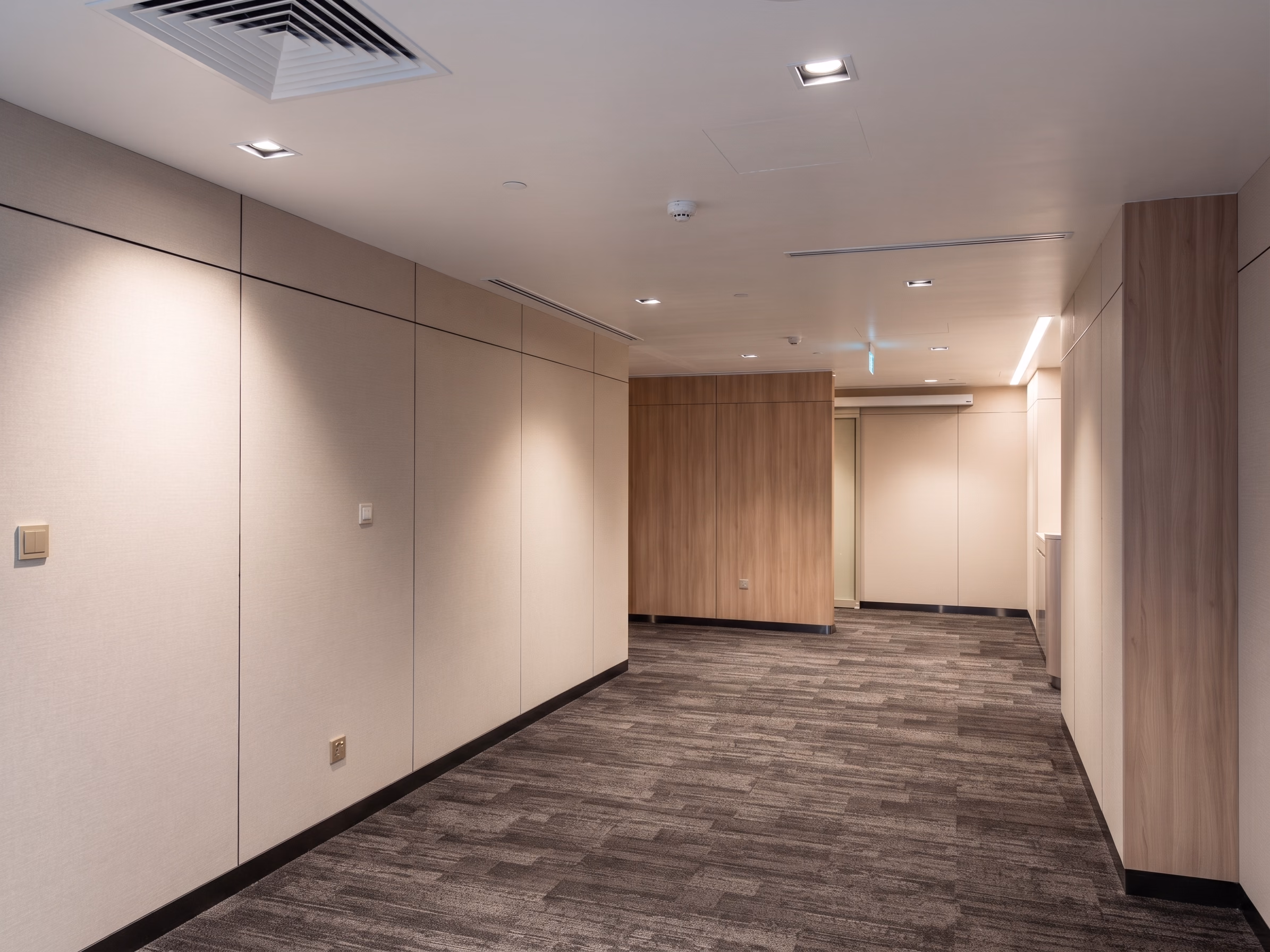 Hospital ward corridor lit by recessed warm downlights, with timber and cream wall panels and no source in the sightline