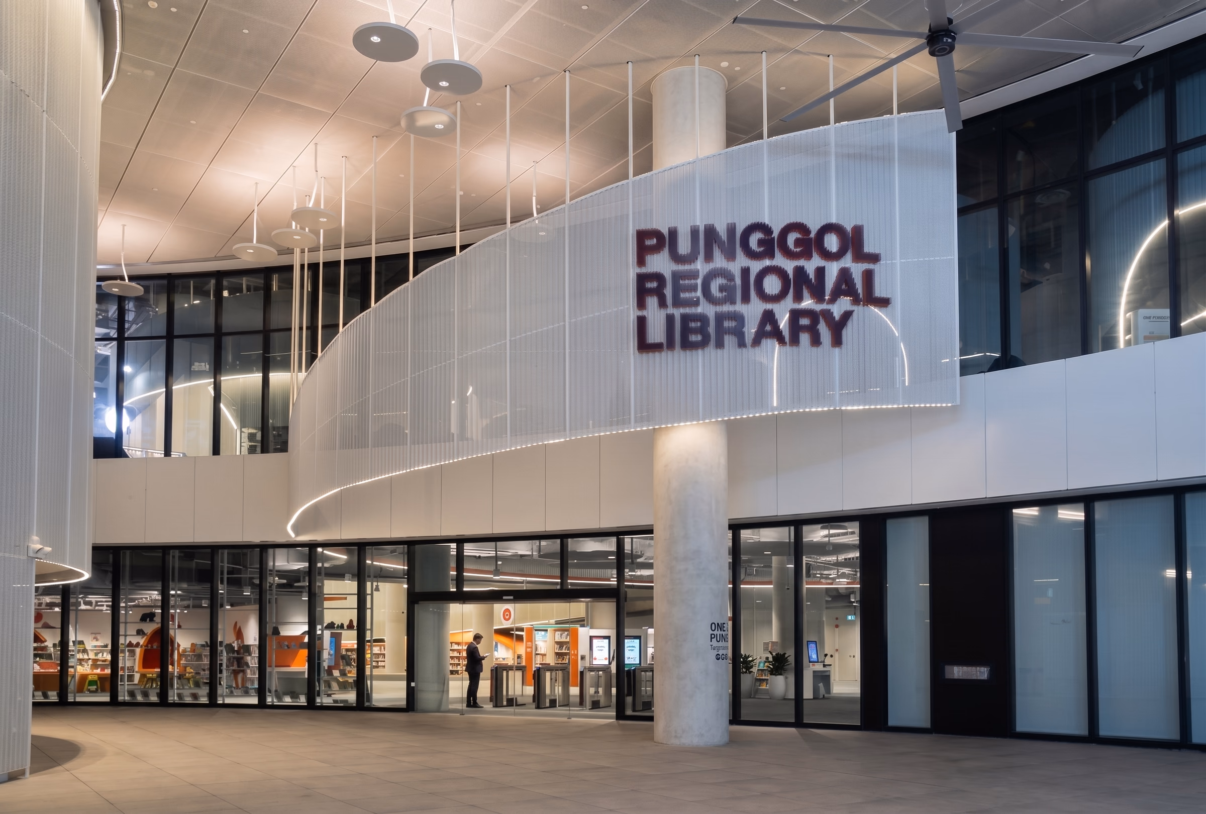 Library entrance at night with a curved perforated signage drum lit from within, above a glazed frontage and warm-lit foyer