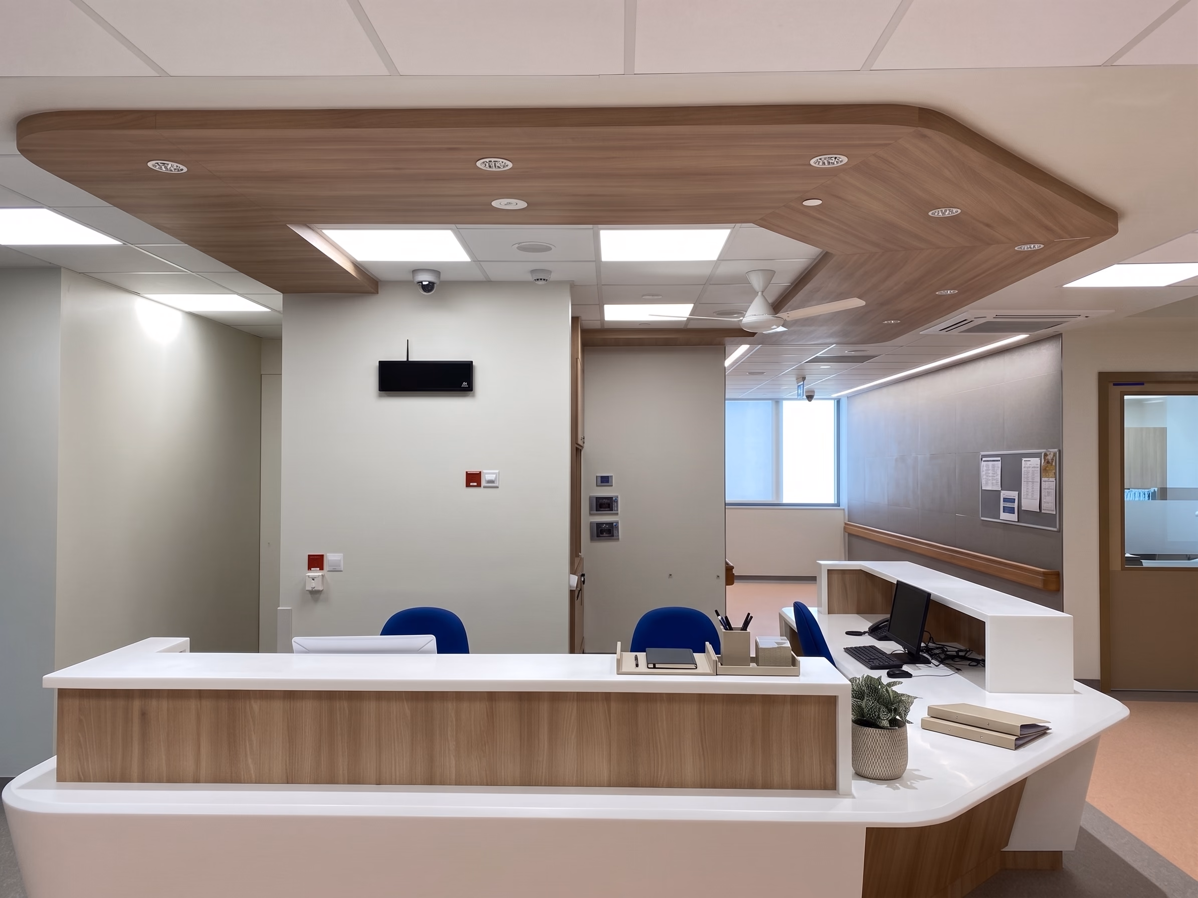 Hospital reception counter under a timber ceiling raft, lit by recessed downlights with even, shadow-free light across the work surface