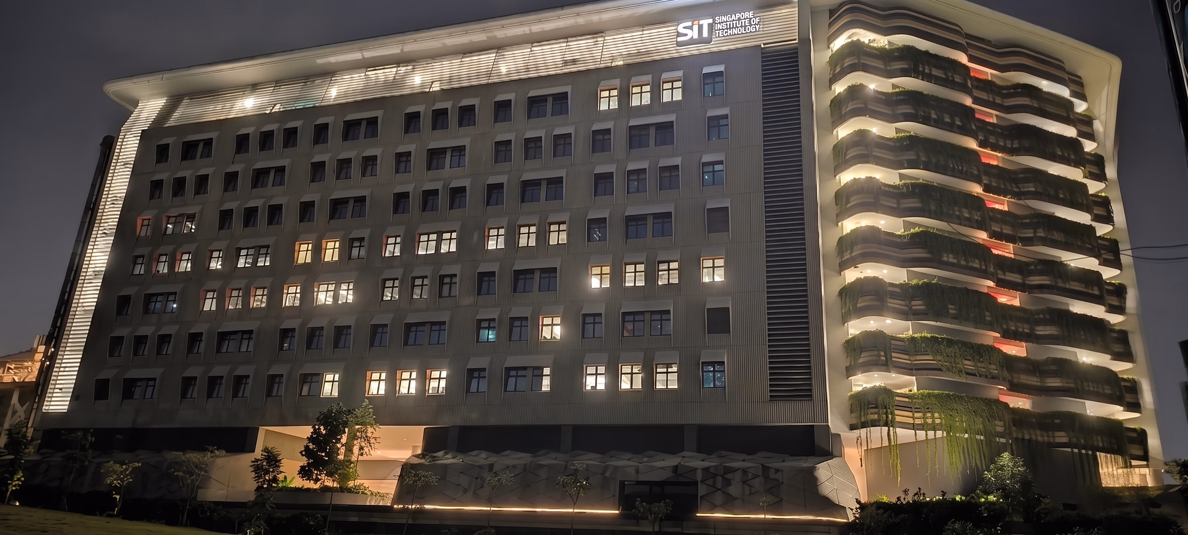 Campus building at night with a grazed flat elevation on one side and stacked planted terraces lit from within on the other